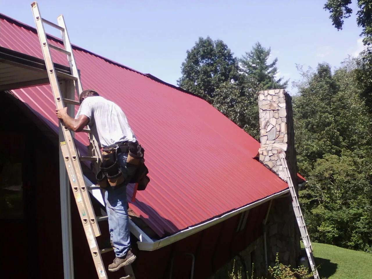 Roofer on a ladder beside a red metal roof and stone chimney — Soffit & Fascia Repair by Roofing Brothers in Bonner